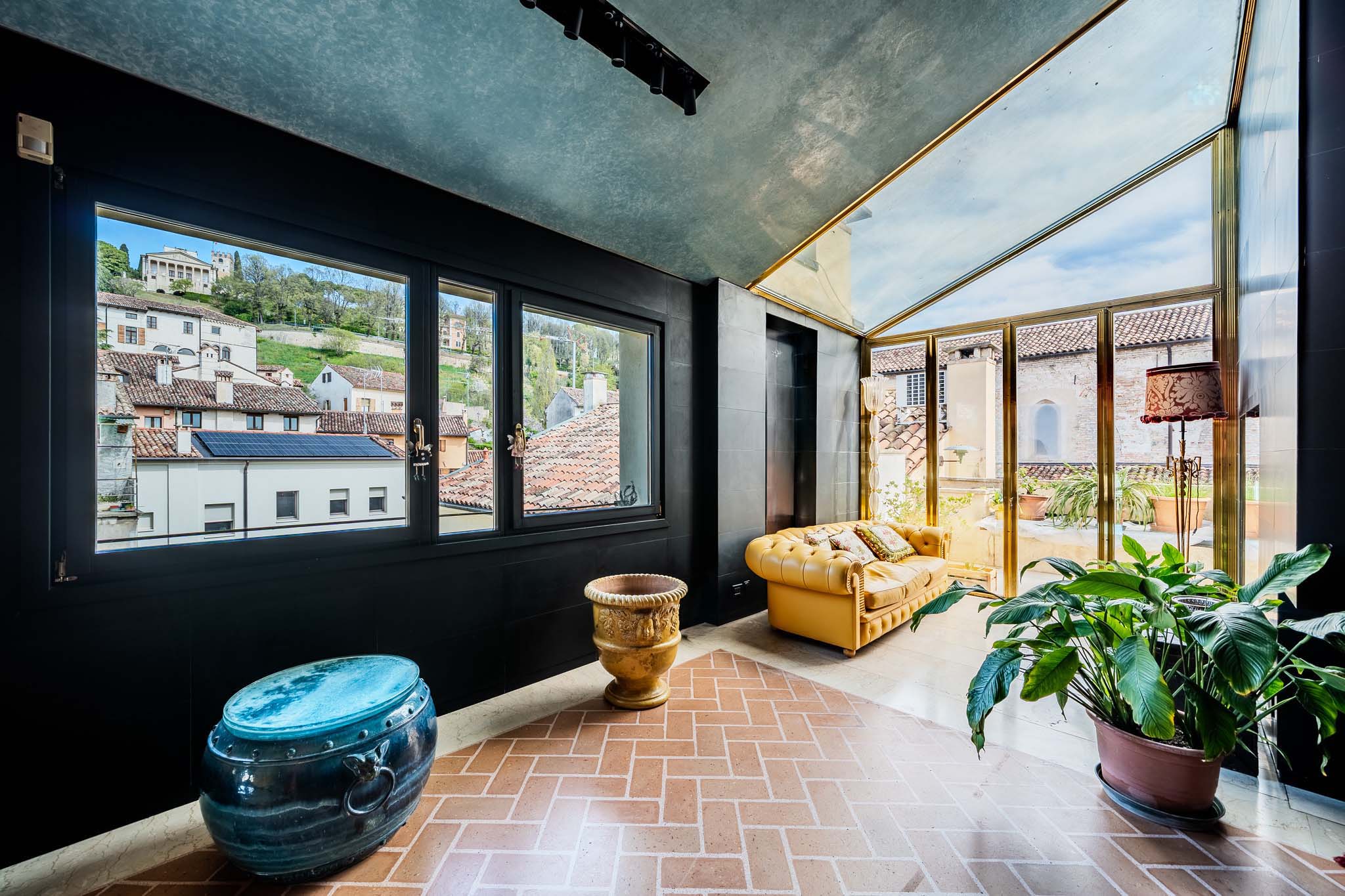 A sunroom with brick floors, a yellow couch, and a view of a European town through a window.