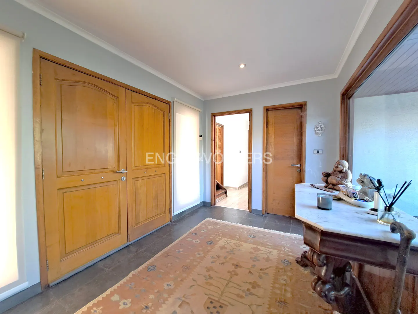 A foyer with light blue walls, a large wooden double door, and a patterned rug on a gray tile floor. A marble-topped table with decorative items sits near a window.