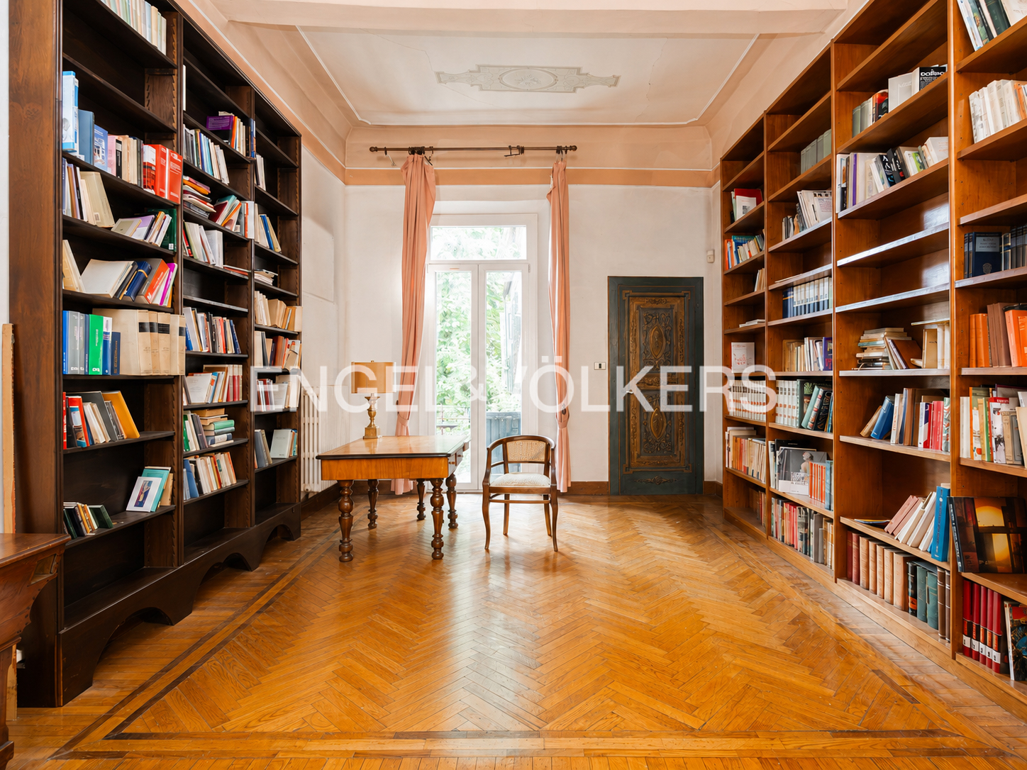 A home library with tall wooden bookshelves, a desk, a chair, and a glass door with pink curtains on a herringbone floor.