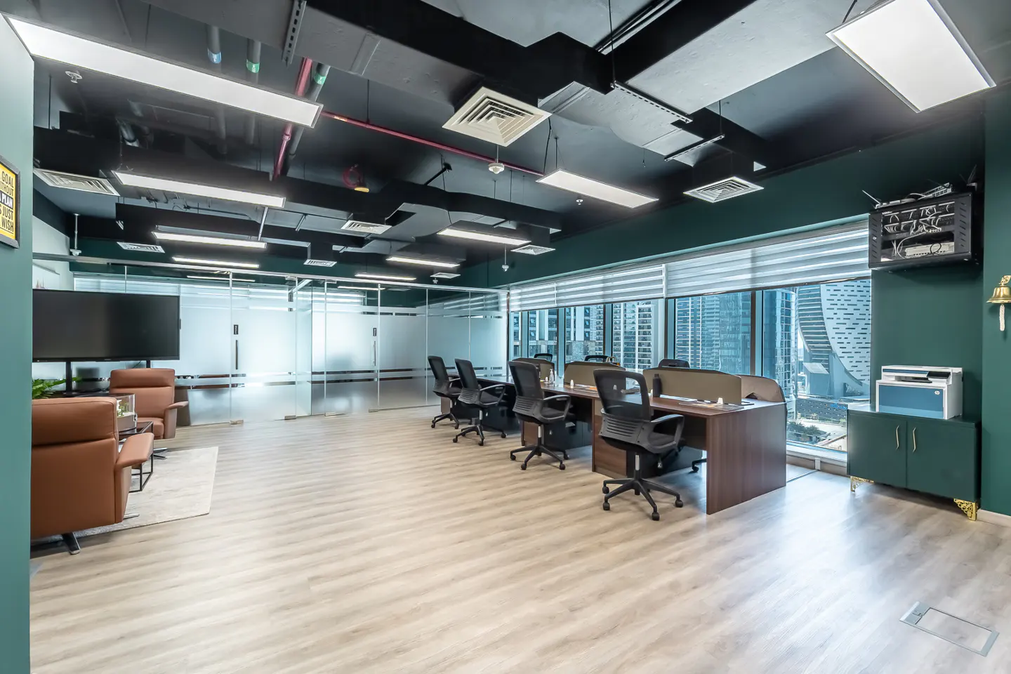 Modern office space with wood floors, black ceiling, and green walls. Desks with black chairs face large windows with a city view. A lounge area is on the left.