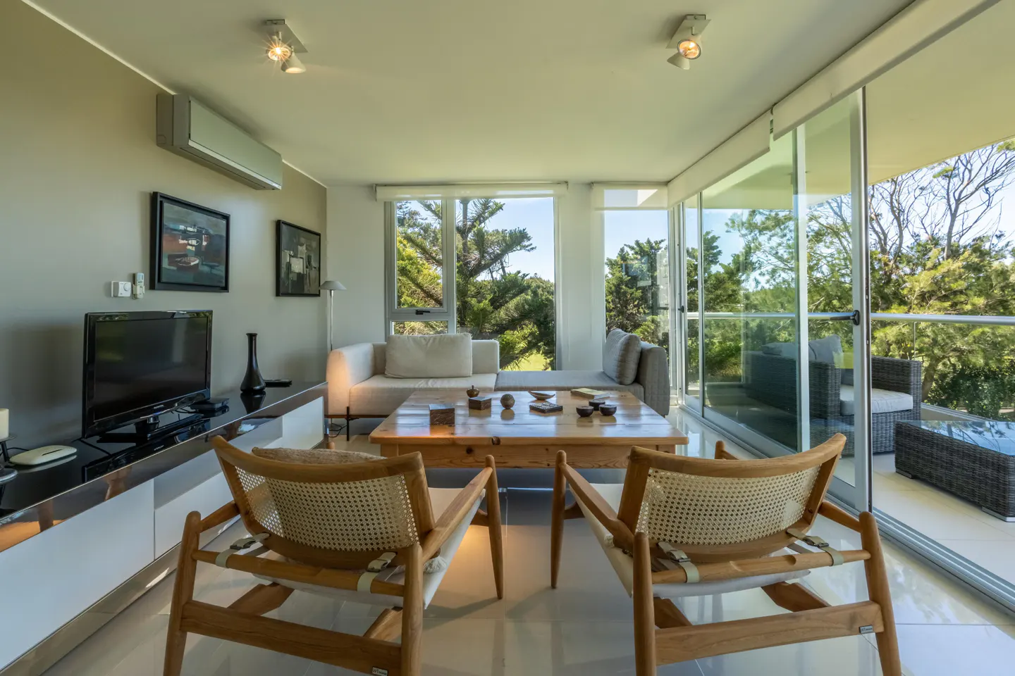 Modern living room with two wooden cane chairs, a coffee table, a white sofa, and a balcony view of trees through glass doors.