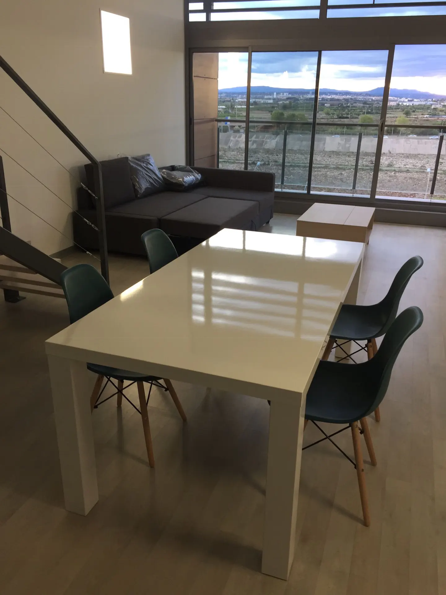 Bright, modern apartment interior with a white table, green chairs, and a gray sofa near a large window with a scenic view.