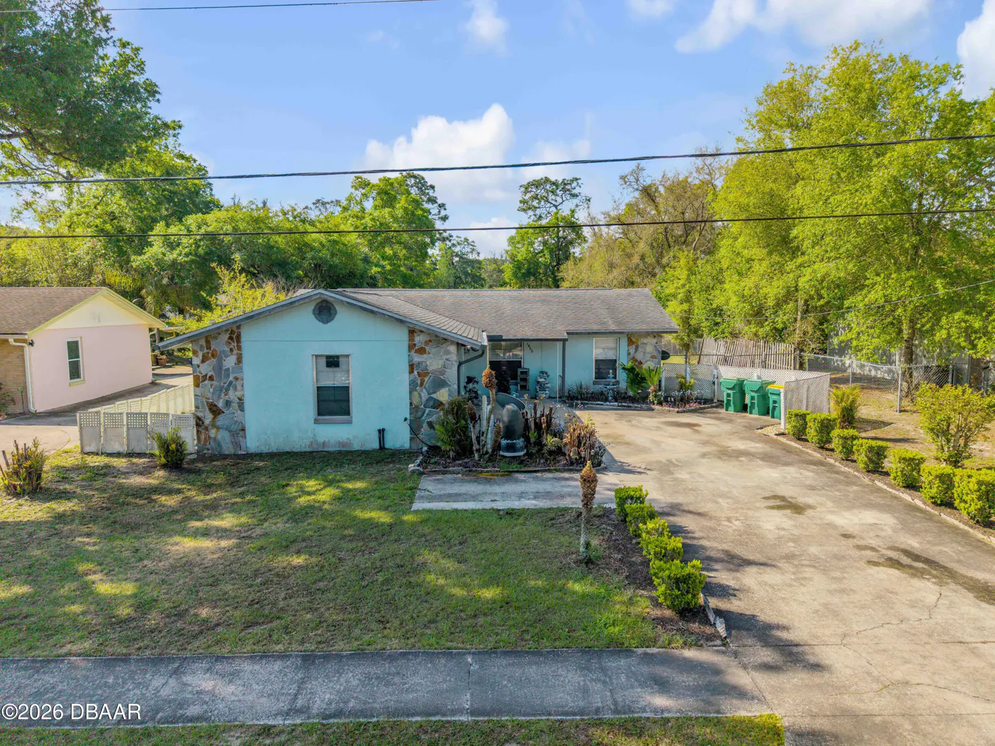A single-story light blue house with stone accents, a gray roof, and a long driveway. Green trees surround the property.