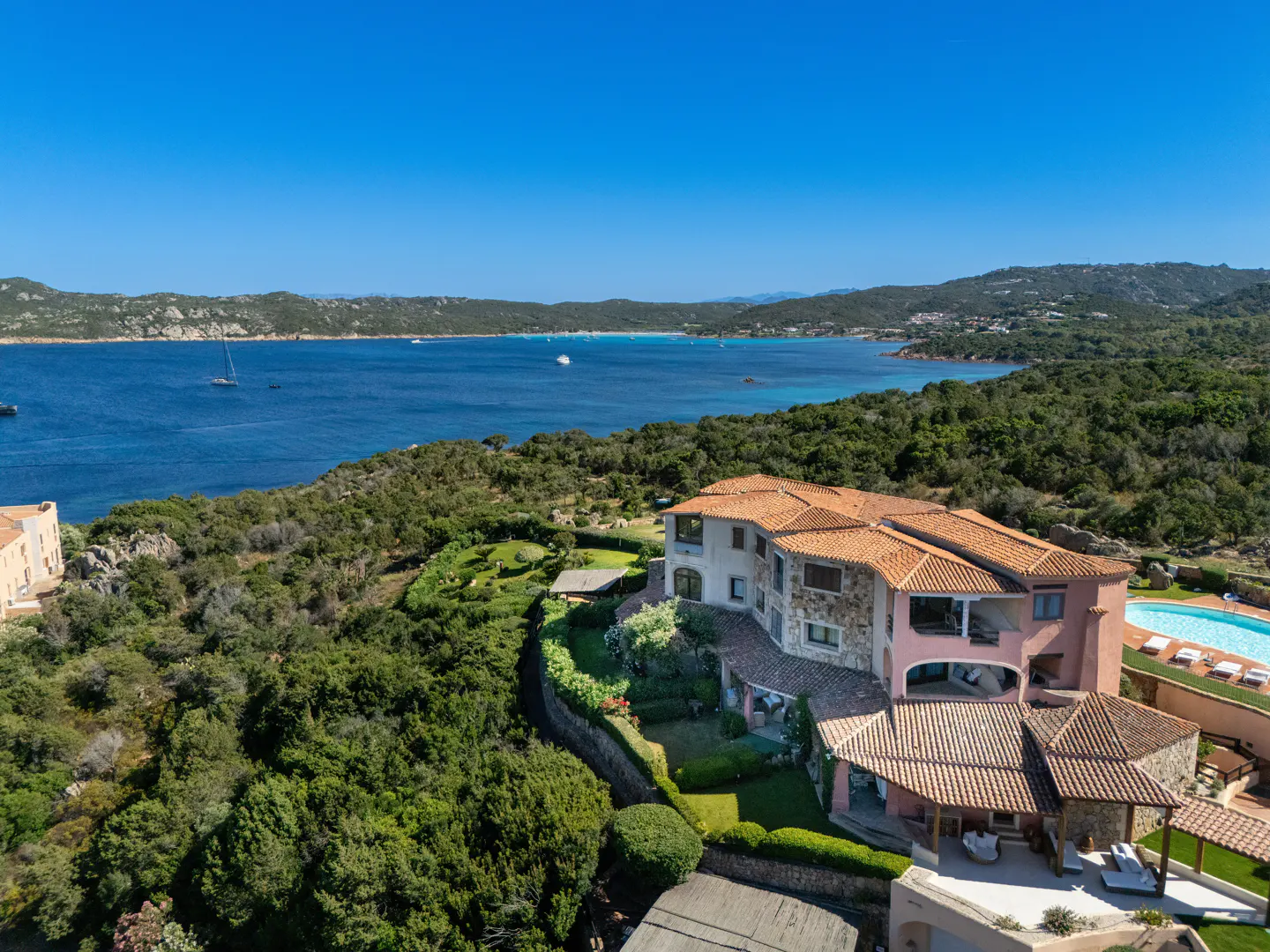 Aerial view of a pink Mediterranean villa with a tiled roof, pool, and lush greenery overlooking a blue bay and hills.