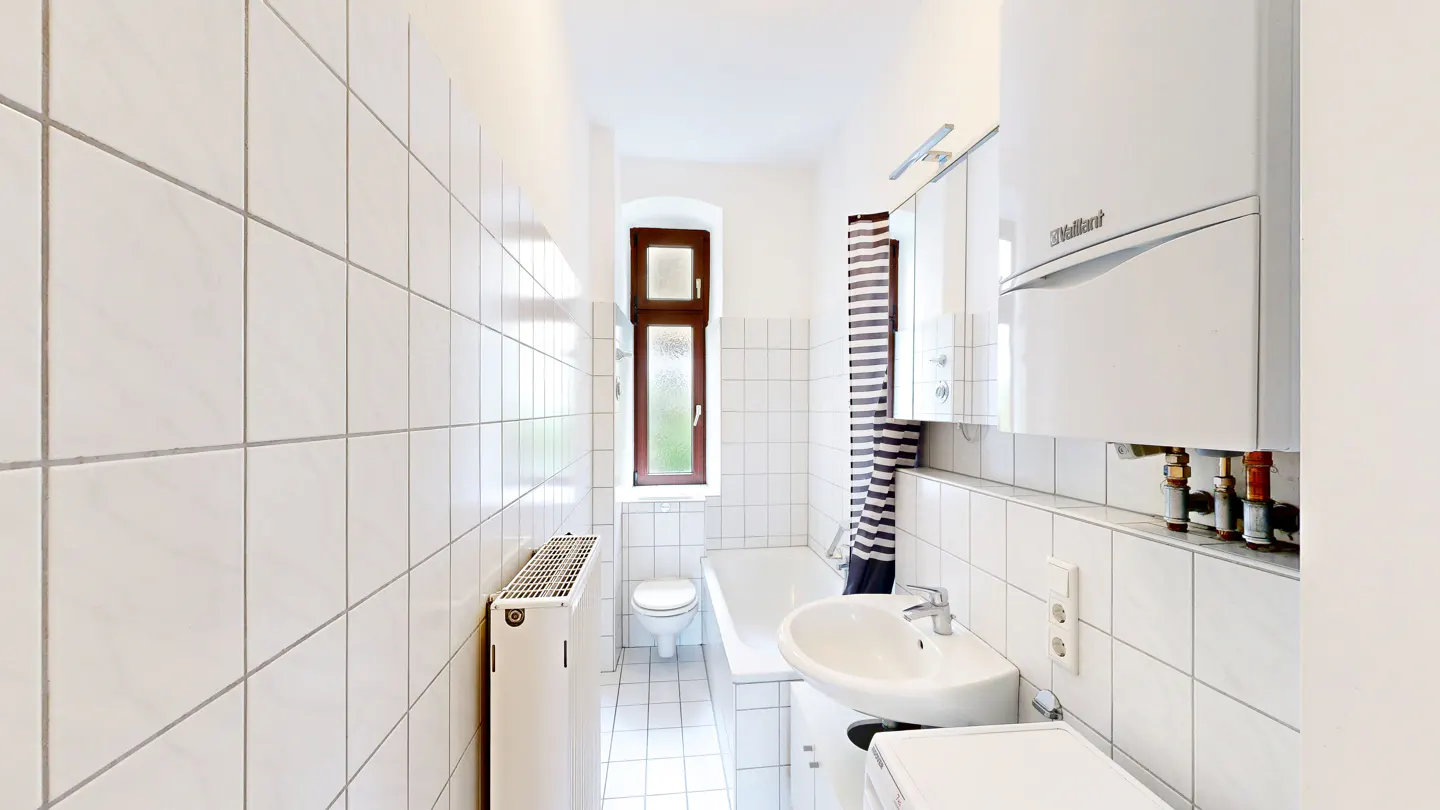 Bright, narrow bathroom with white square tiles, a window, a sink, and a white bathtub. A striped shower curtain hangs near the tub.