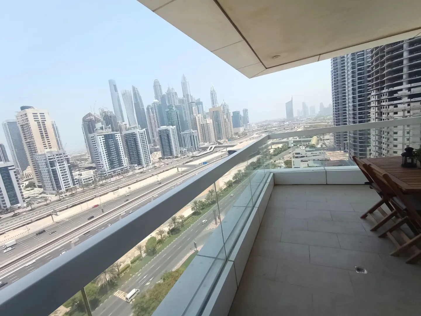 Balcony view of Dubai skyline. Glass railing overlooks a busy highway and city buildings. Wooden table and chairs on the tiled balcony.