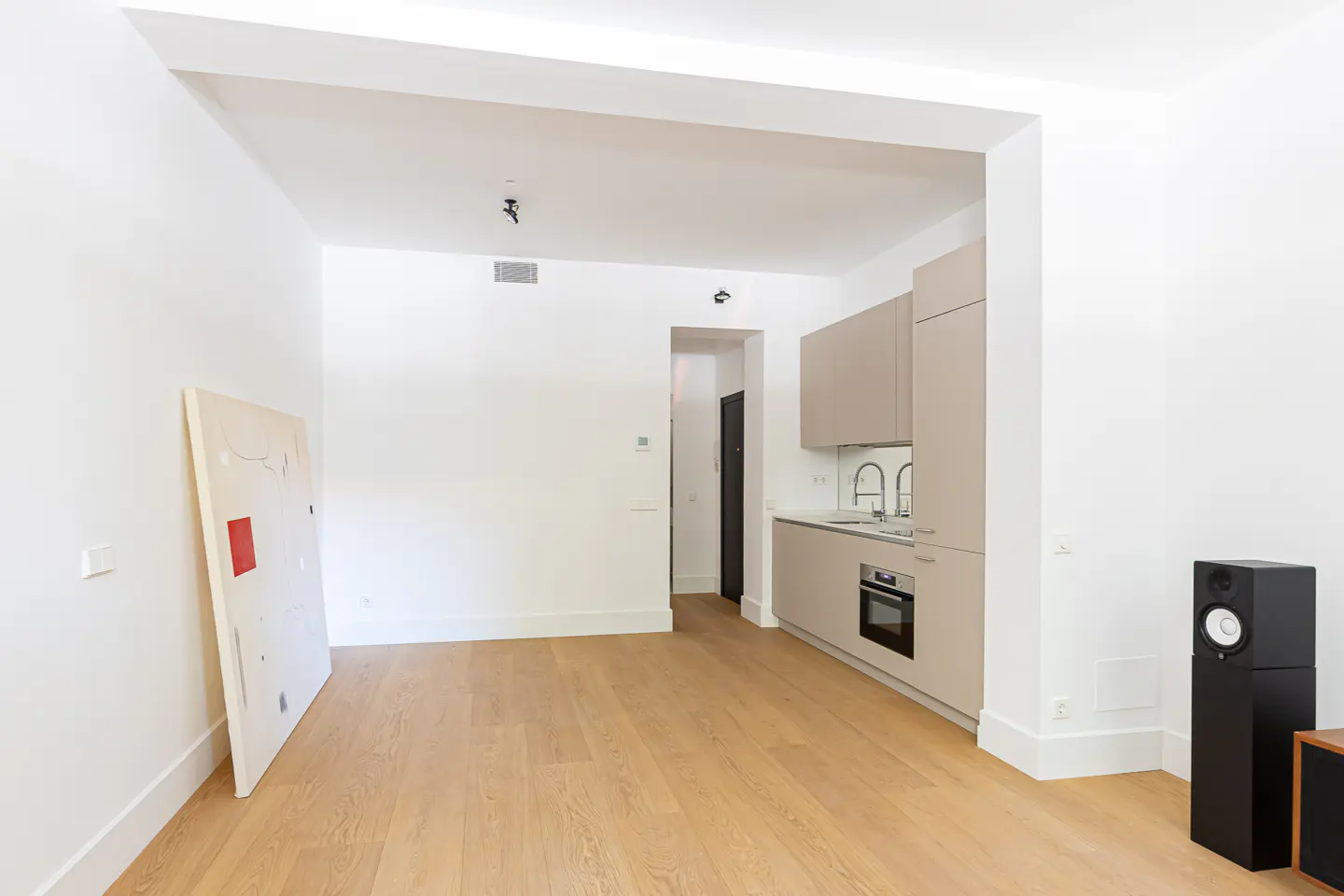 Minimalist living area with light wood floors, a modern beige kitchenette, and a large abstract painting leaning on a wall.