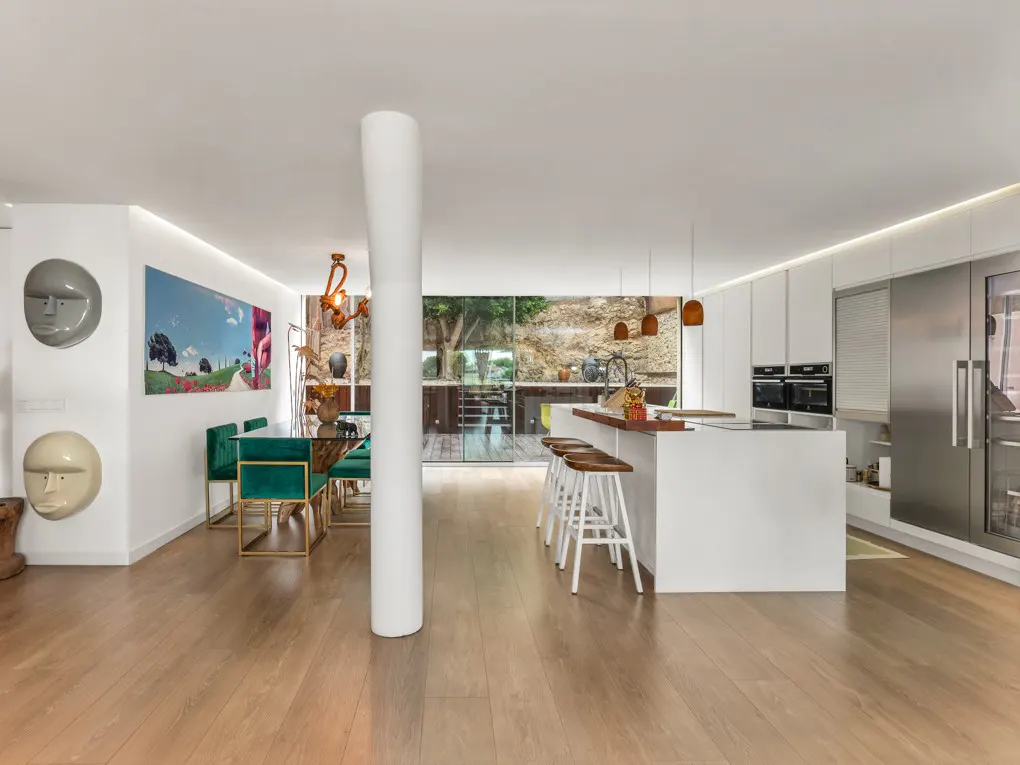 Open-concept kitchen and dining area with wood floors, white walls, and modern decor. Emerald chairs surround a glass table. A white island has stools.
