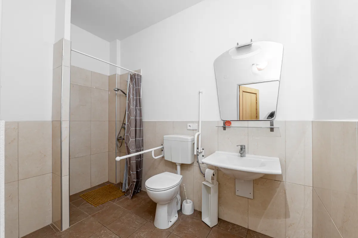 Accessible bathroom with white toilet, sink, and grab bars. Shower with gray curtain, beige tile, and brown floor. Mirror above sink.