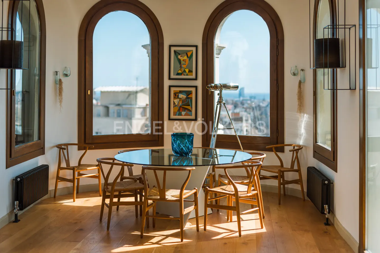 Bright dining room with wood floors, arched windows, and city views. A glass table is surrounded by wooden chairs. A telescope stands near the window.