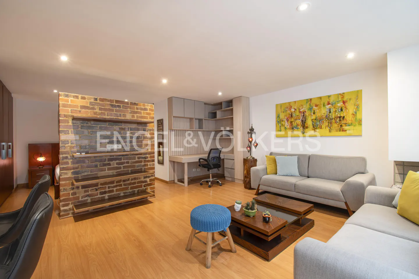 Bright living room with wood floors, gray sofas, and a brick accent wall. A desk area is visible in the background.