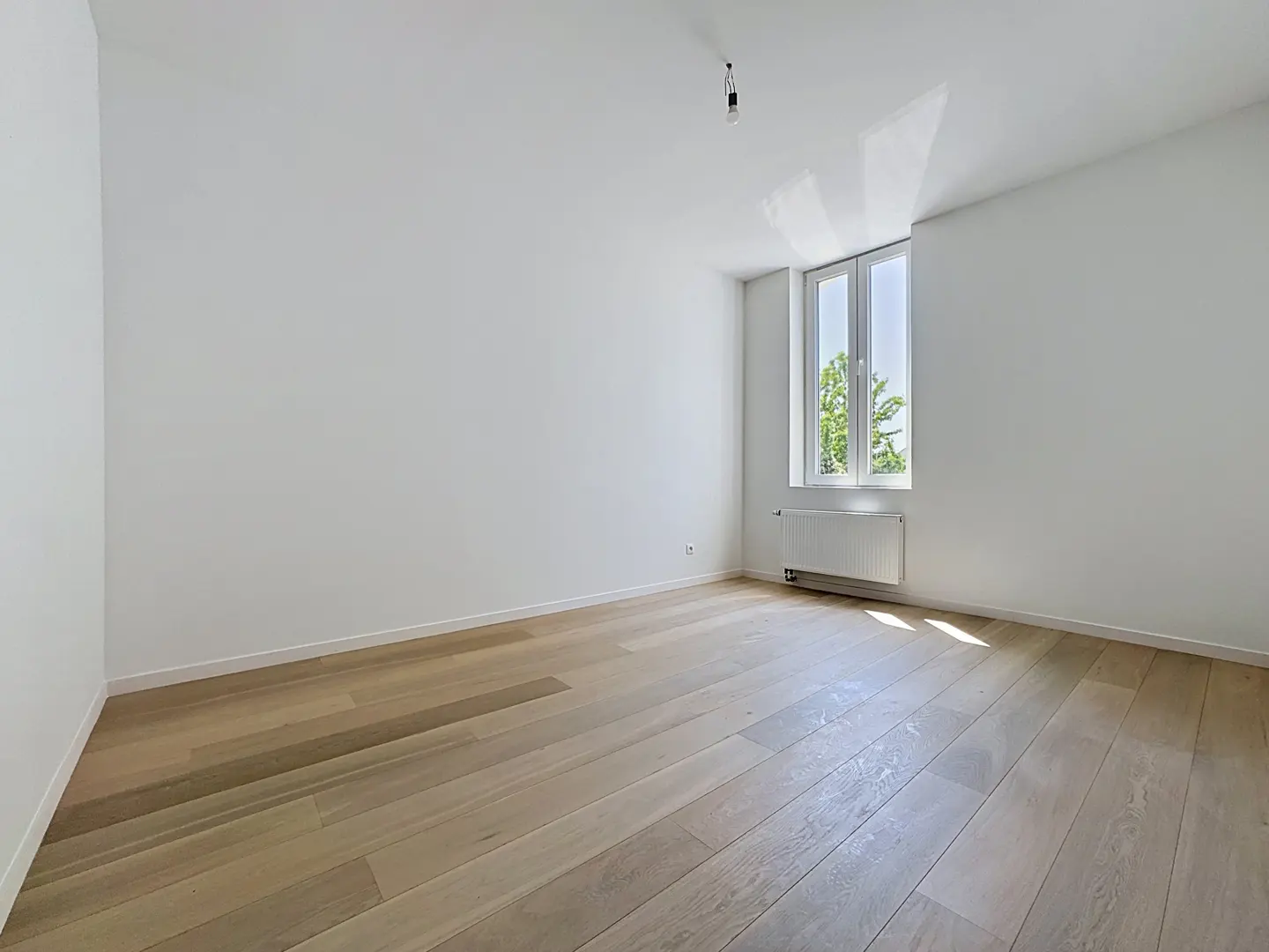 Empty room with white walls, light wood flooring, a single window, and a radiator under the window.
