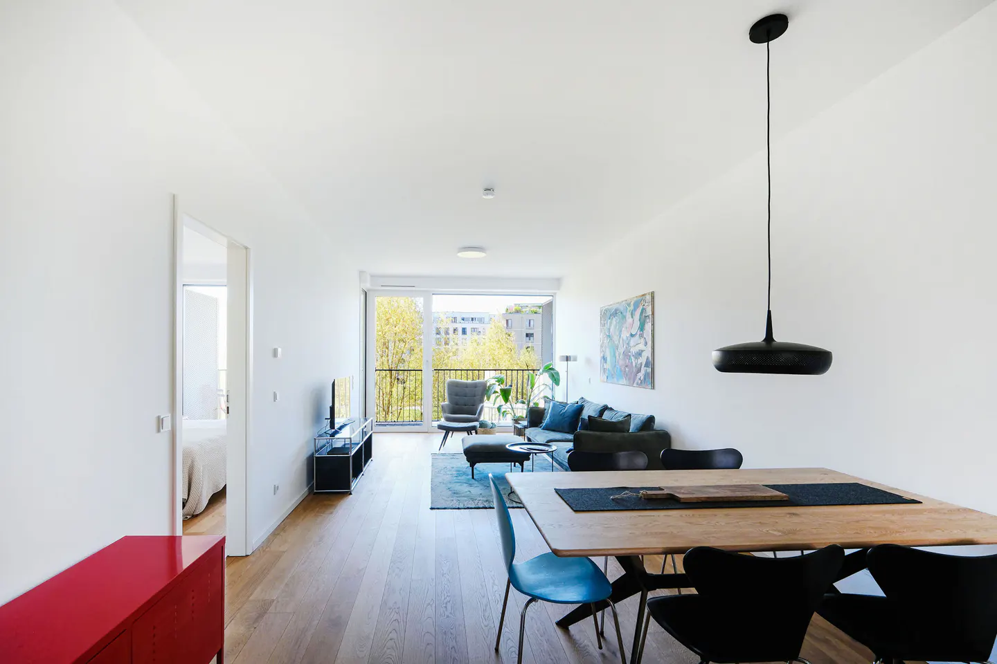 Bright, modern apartment interior with wood floors, white walls, dining table, and living area with balcony view.