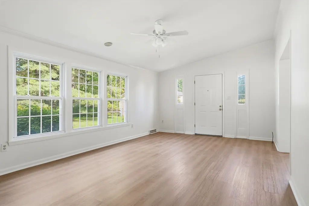 Bright, empty living room with light wood floors, white walls, and a white front door. Three large windows overlook a green yard. A white ceiling fan is centered.