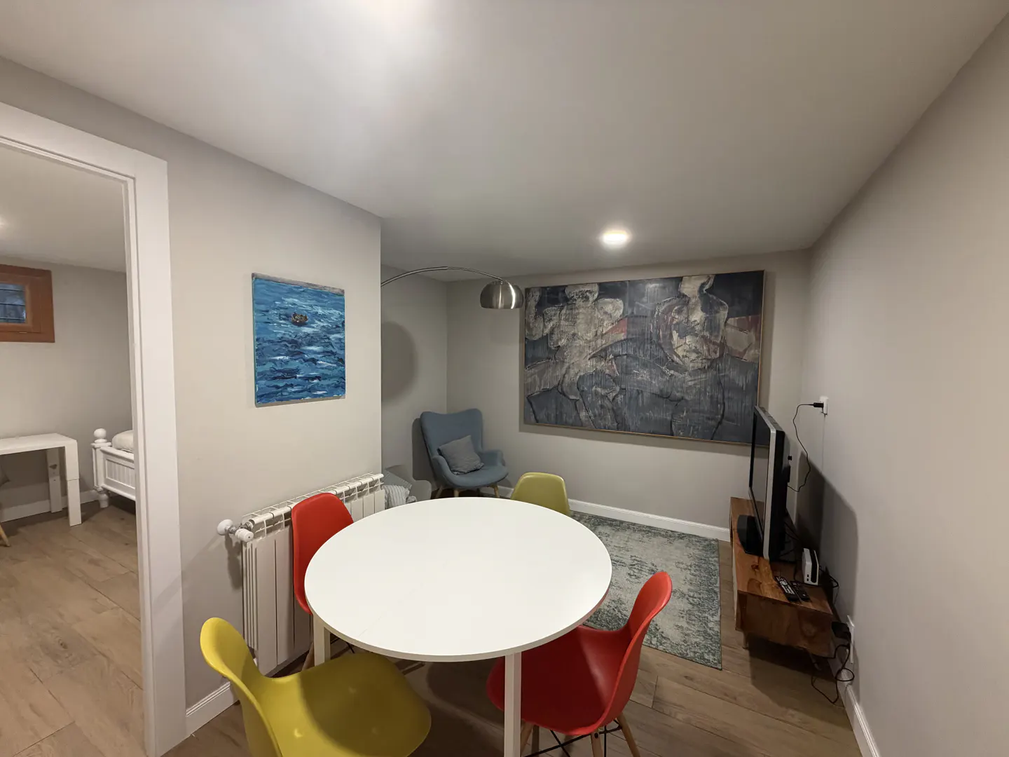 Living room with a white round table, red and yellow chairs, and abstract art on the walls. A TV sits on a wooden stand.
