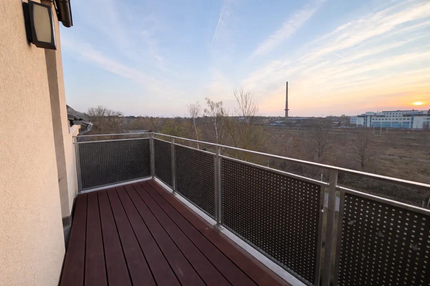 Balcony with dark wood flooring and metal railings overlooking a sunset landscape with a distant industrial chimney.
