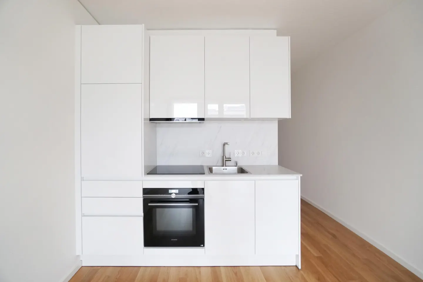 A compact, modern kitchen with white cabinets, a black oven, a stainless steel sink, and light wood flooring.