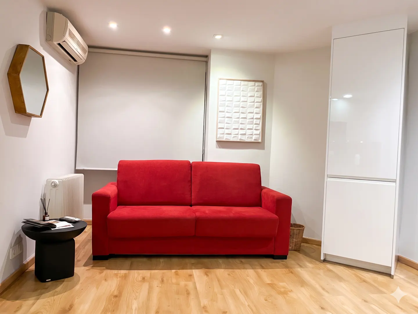 A living room with a red sofa, white walls, and light wood floors. A black side table and white storage unit are also visible.