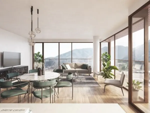 Bright, modern living room with floor-to-ceiling windows showing mountain views. Green chairs surround a marble table. A gray sofa sits in the background.