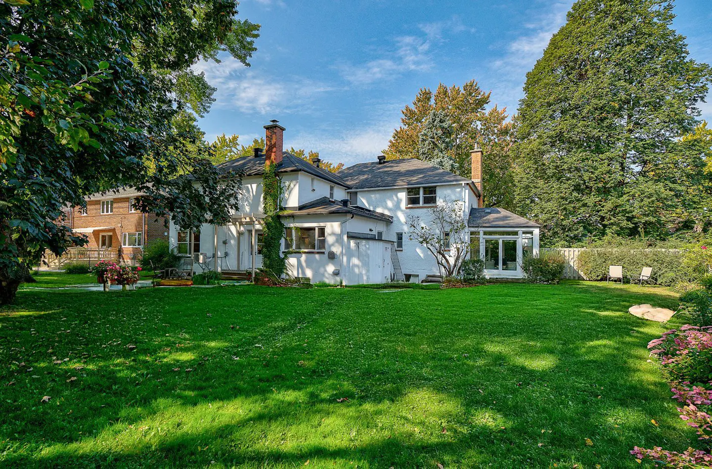 Backyard view of a white house with a large green lawn, trees, and blue sky.