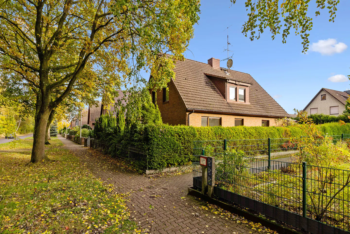A brick house with a brown roof is surrounded by green hedges and a black metal fence on a sunny autumn day.