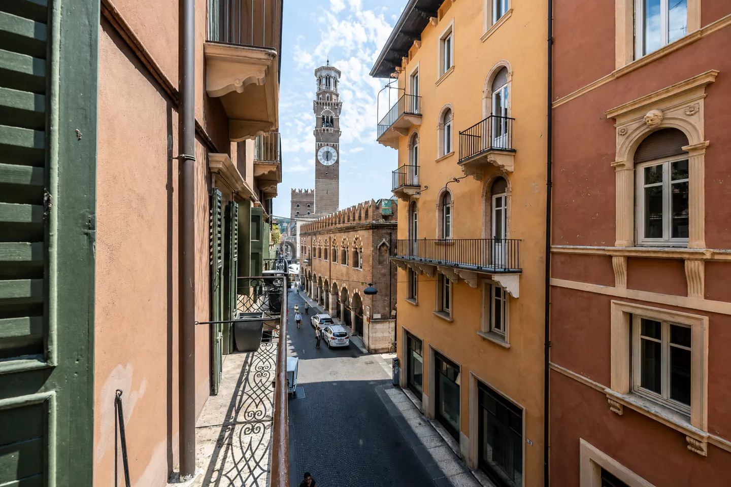 View of Verona street with tall tower. Buildings are peach, yellow, and red. Balconies and green shutters are visible.
