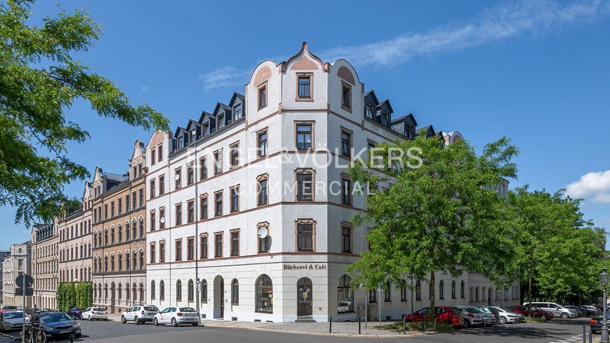A white corner apartment building with a ground-floor bakery and cafe, set on a sunny street with parked cars and trees.