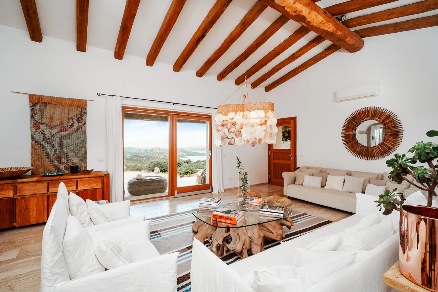 Bright living room with white sofas, wood beams, and a glass coffee table. Doors open to a scenic view.