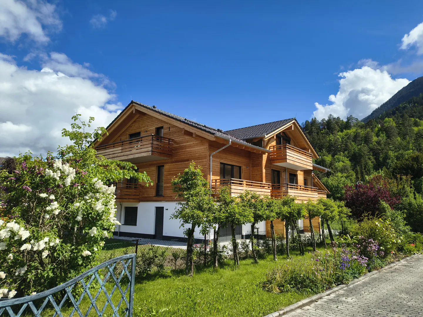 Two-story wood house with balconies, set against a blue sky and mountain backdrop. Lush greenery surrounds the property.