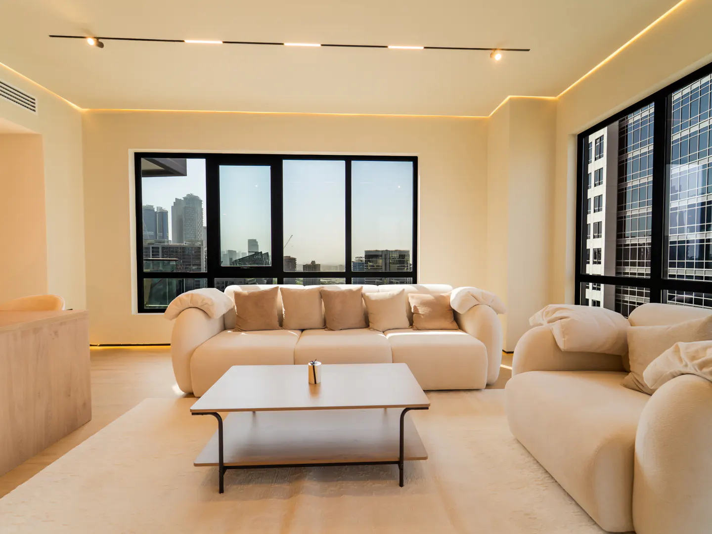 Bright living room with a cream sofa, pillows, and rug. Large windows offer a city view. A modern coffee table sits in the center.