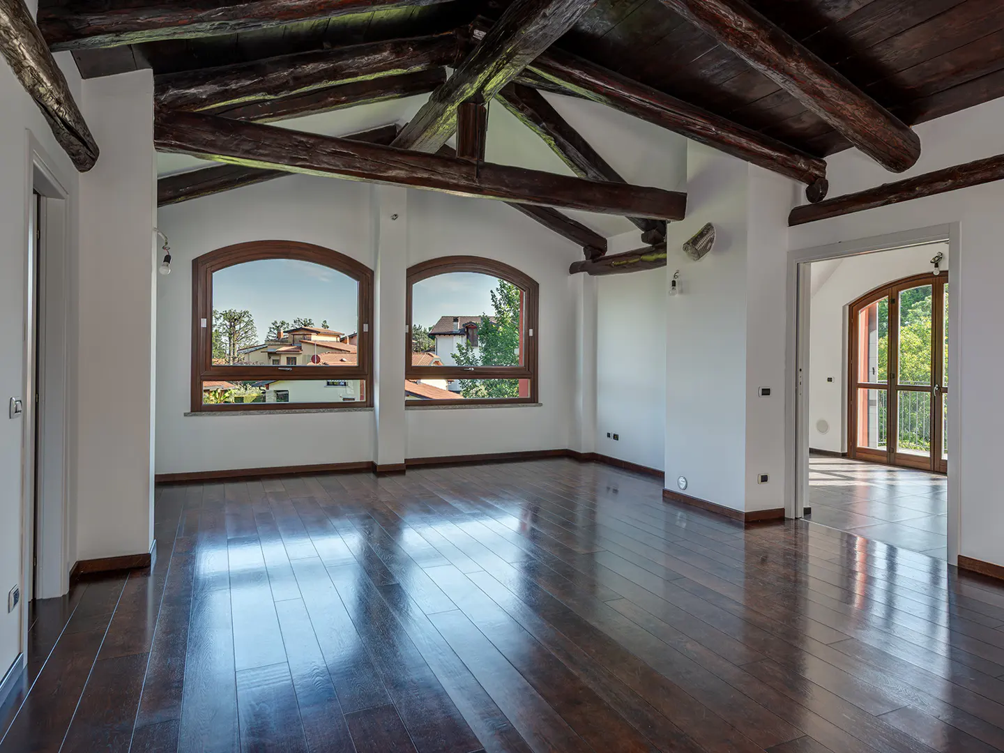 Spacious room with dark wood floors, vaulted ceiling with exposed wooden beams, and arched windows overlooking a neighborhood.