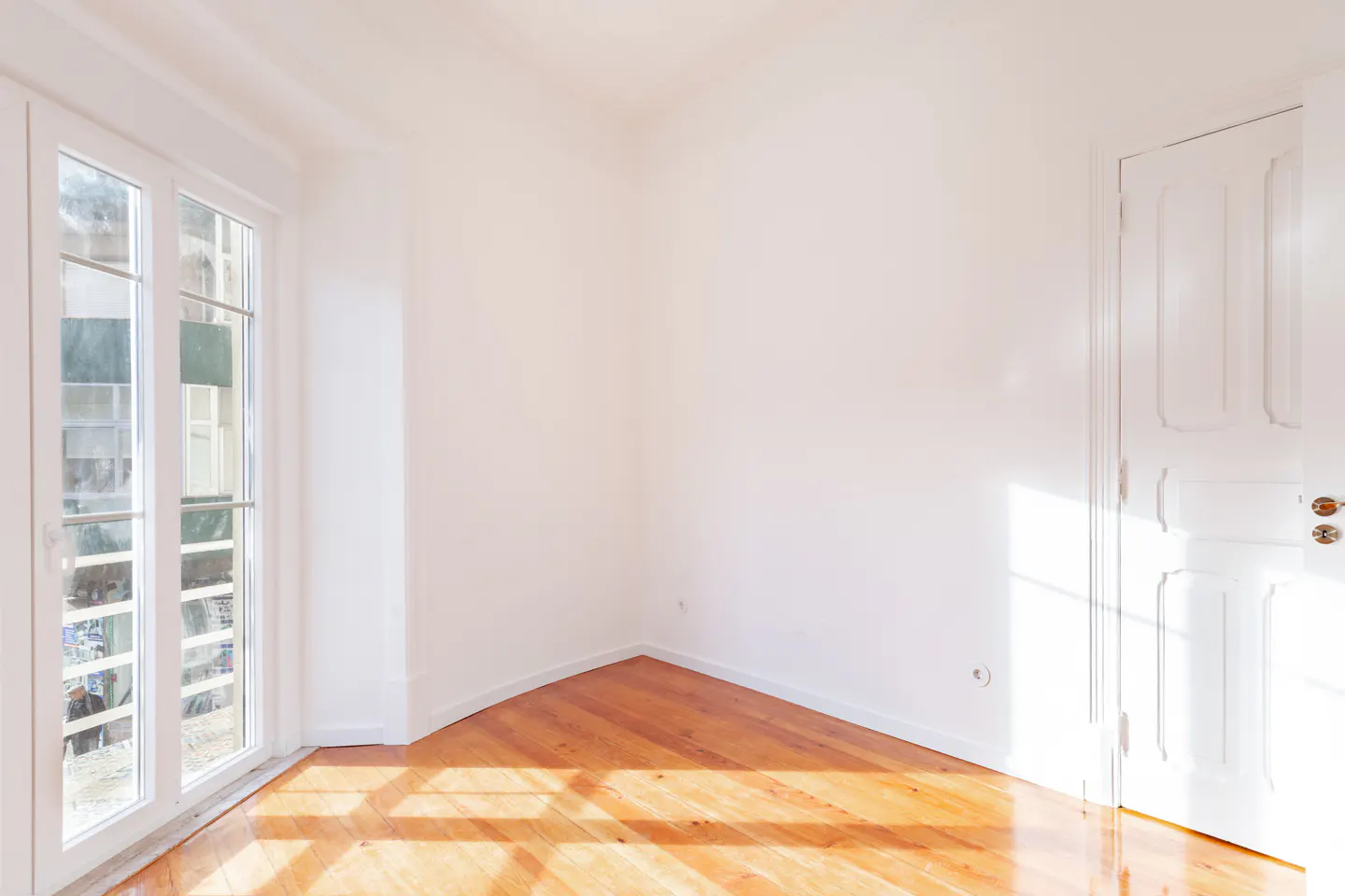 Bright, empty room with white walls, wood floor, and a white door. Sunlight streams in through a multi-paned window.