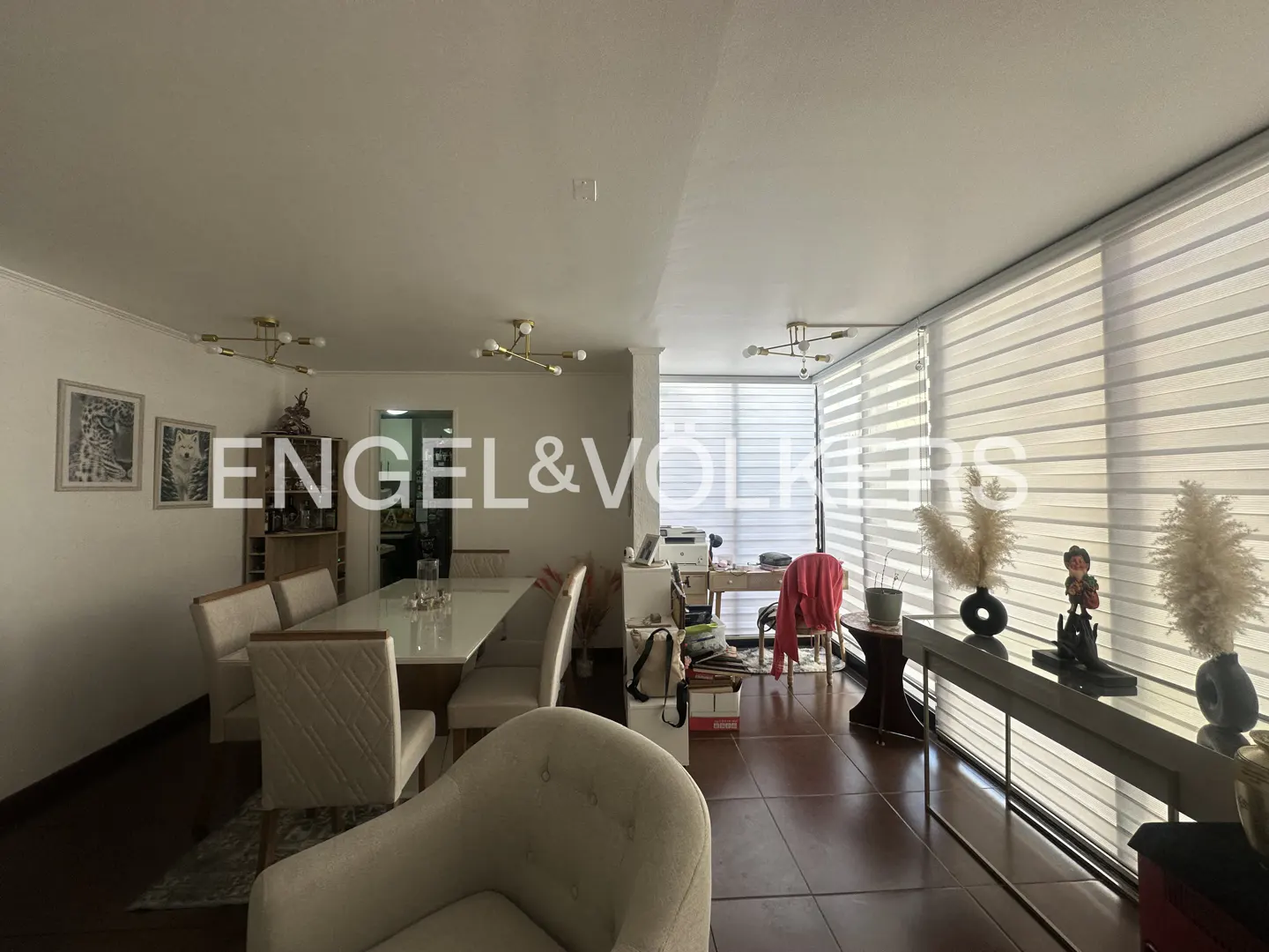 A bright, open-concept living and dining area with a long table, chairs, and large windows with blinds.