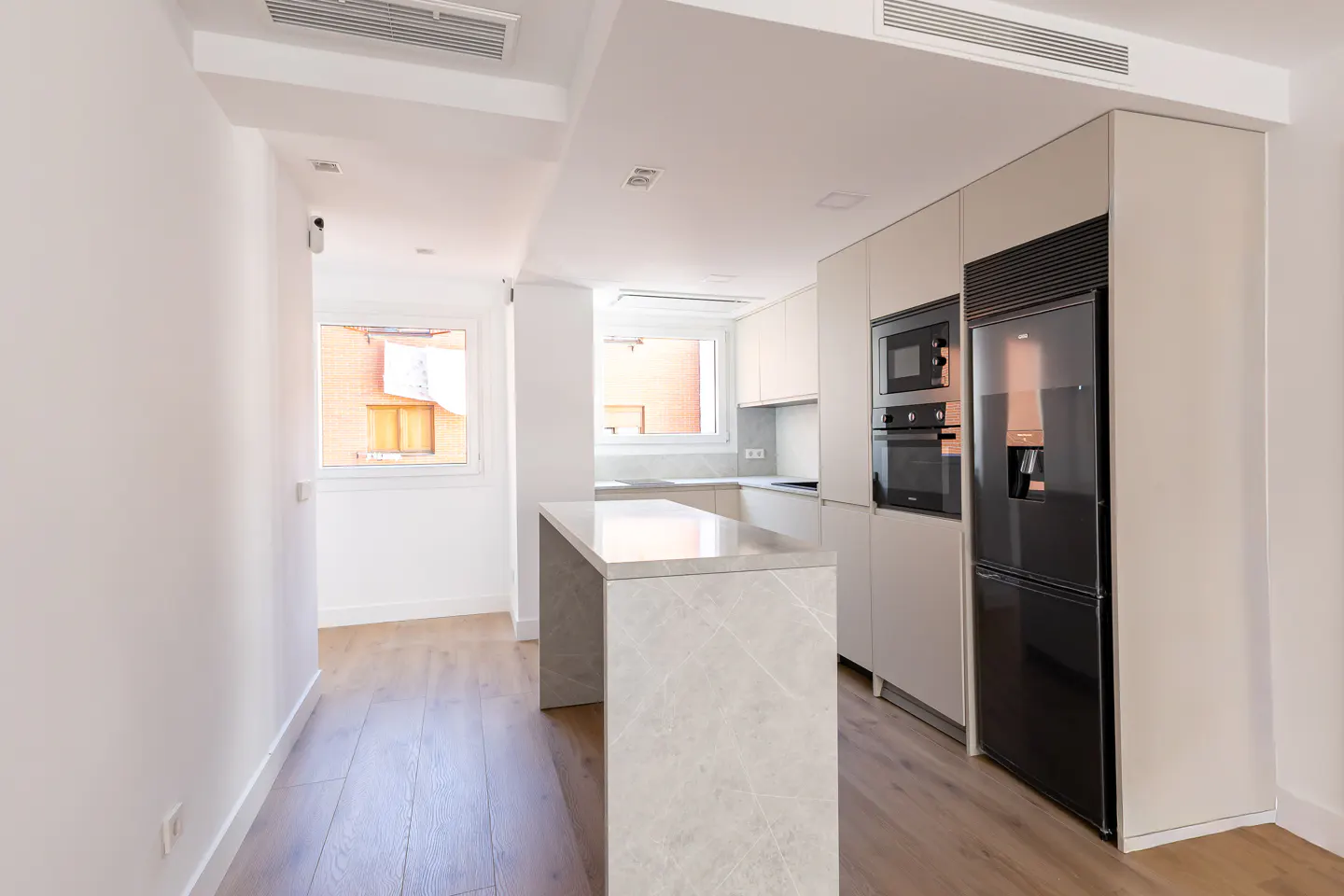 A modern kitchen with white walls, light wood floors, and a marble island. Appliances include a black refrigerator, microwave, and oven. A window provides natural light.