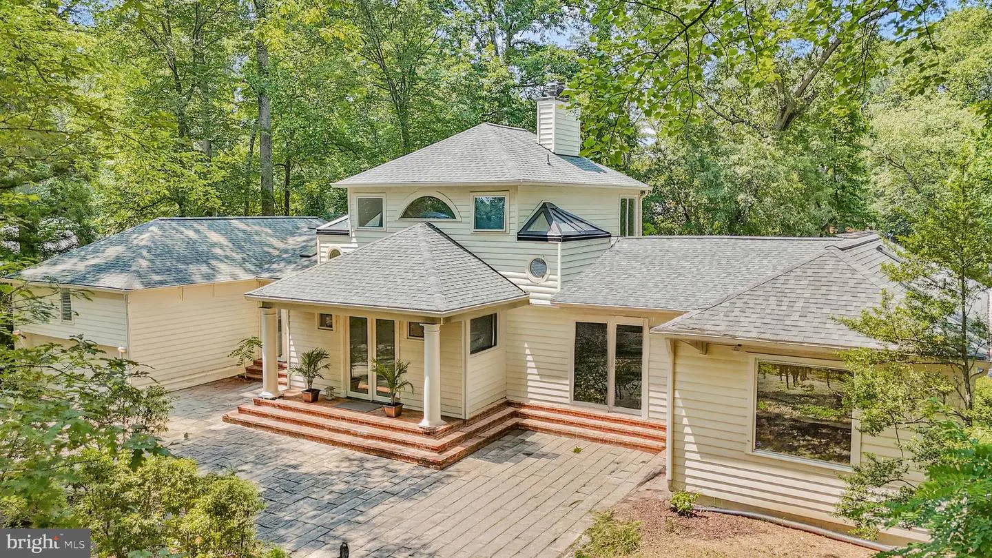 Exterior view of a two-story, light yellow house with a gray roof, surrounded by green trees. A stone patio leads to the front entrance.