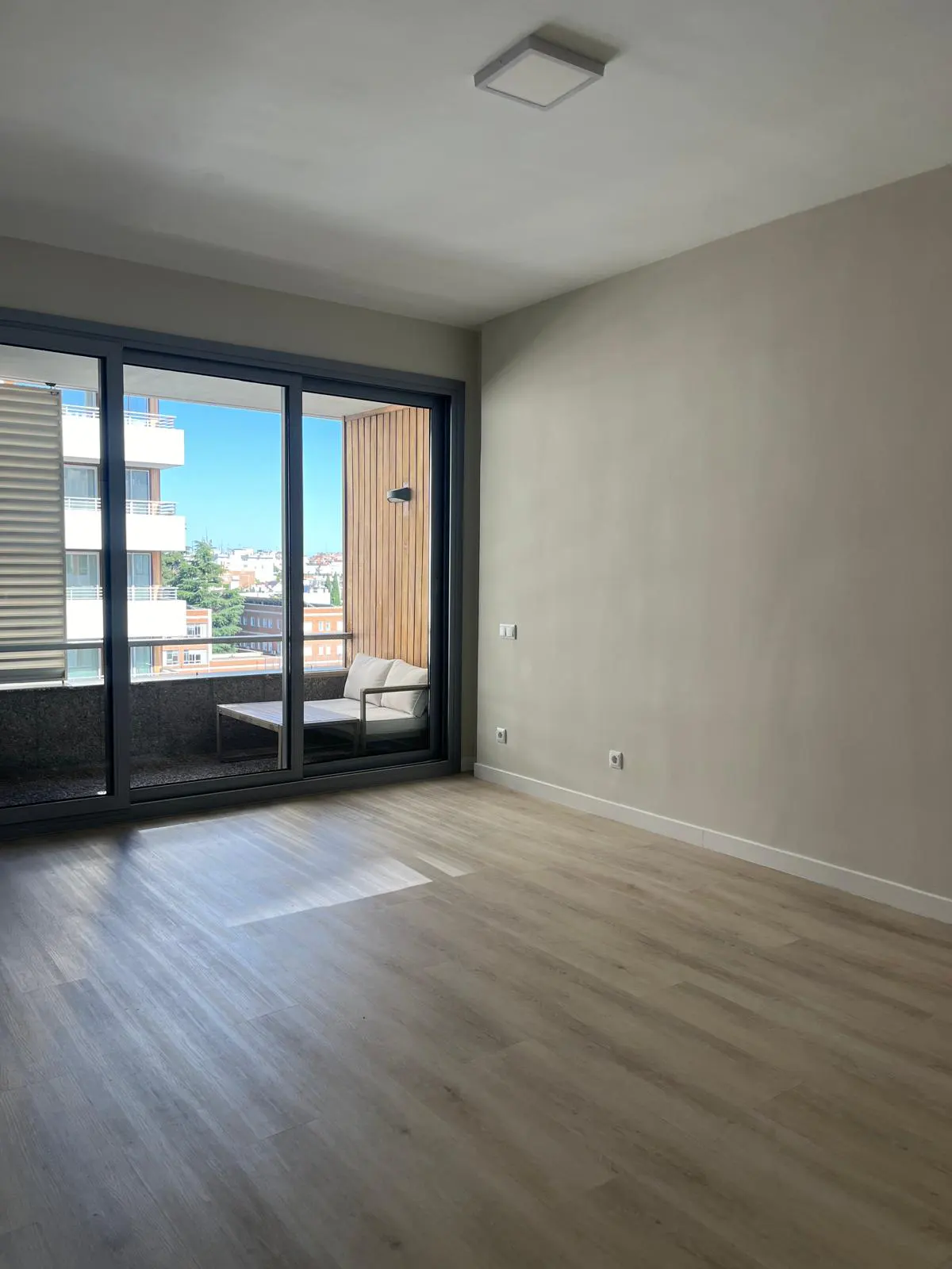 Empty room with light wood floors and beige walls. Sliding glass doors open to a balcony with outdoor seating and a city view.