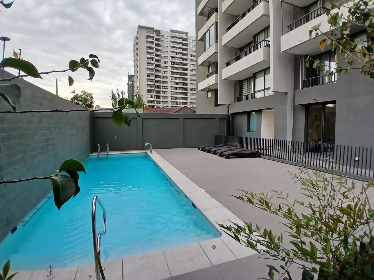 Outdoor pool with blue water, surrounded by gray walls and lounge chairs. Tall apartment buildings are in the background.