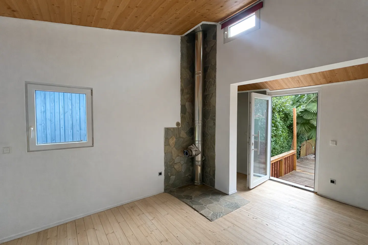Bright room with wood floors, white walls, and wood ceiling. A stone fireplace with a metal flue is centered. Open doors lead to a deck and lush greenery.