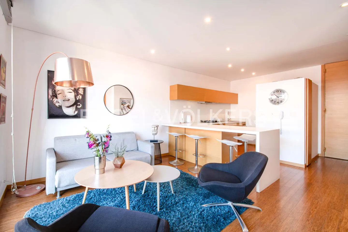 Modern open-plan living room with a grey sofa, blue rug, kitchen island with bar stools, and a tall copper floor lamp.