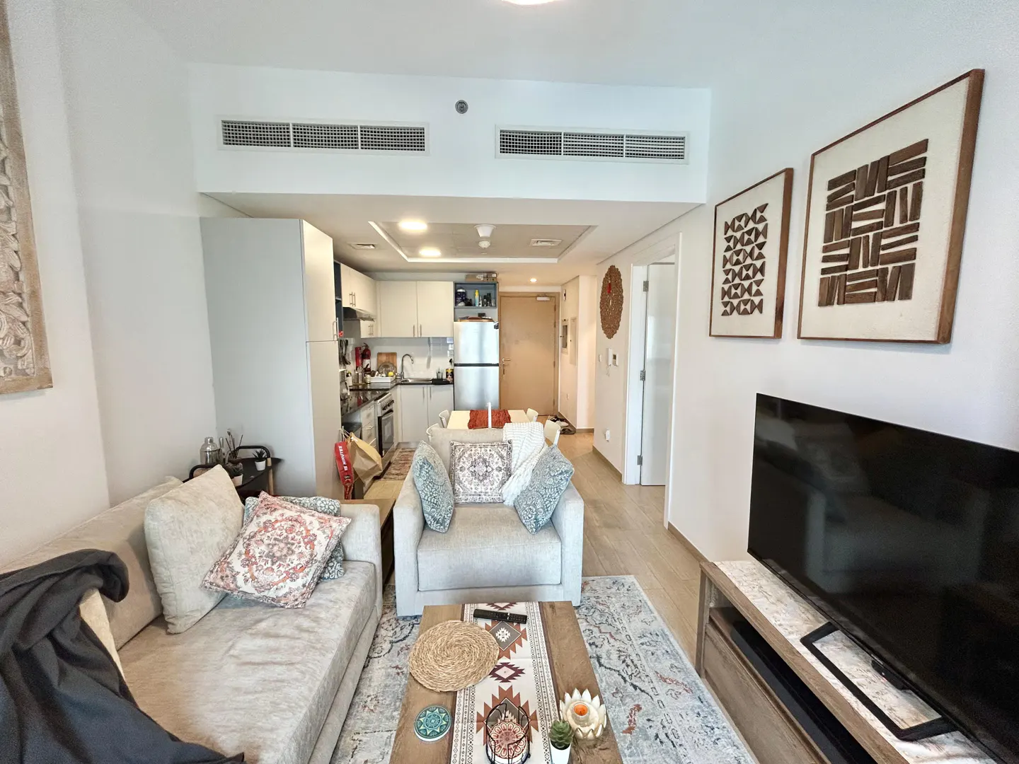 A bright, modern apartment interior with a sofa, armchair, TV, and open kitchen. Artwork adorns the white walls.