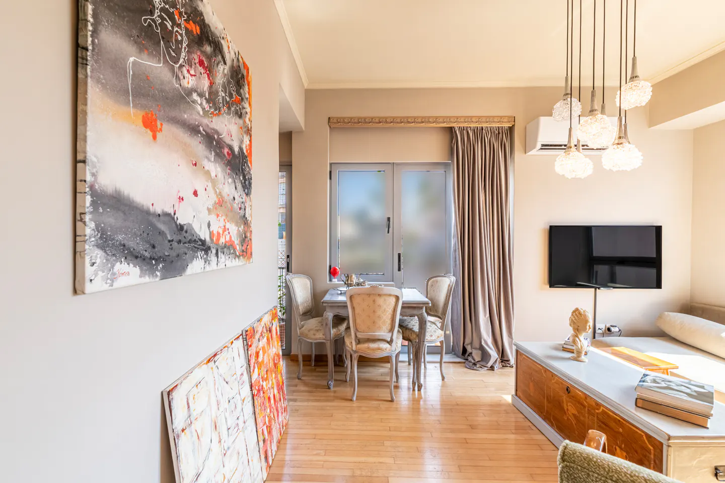 Modern living room with hardwood floors, abstract art, a dining set by a window, and a wall-mounted TV above a wooden chest.