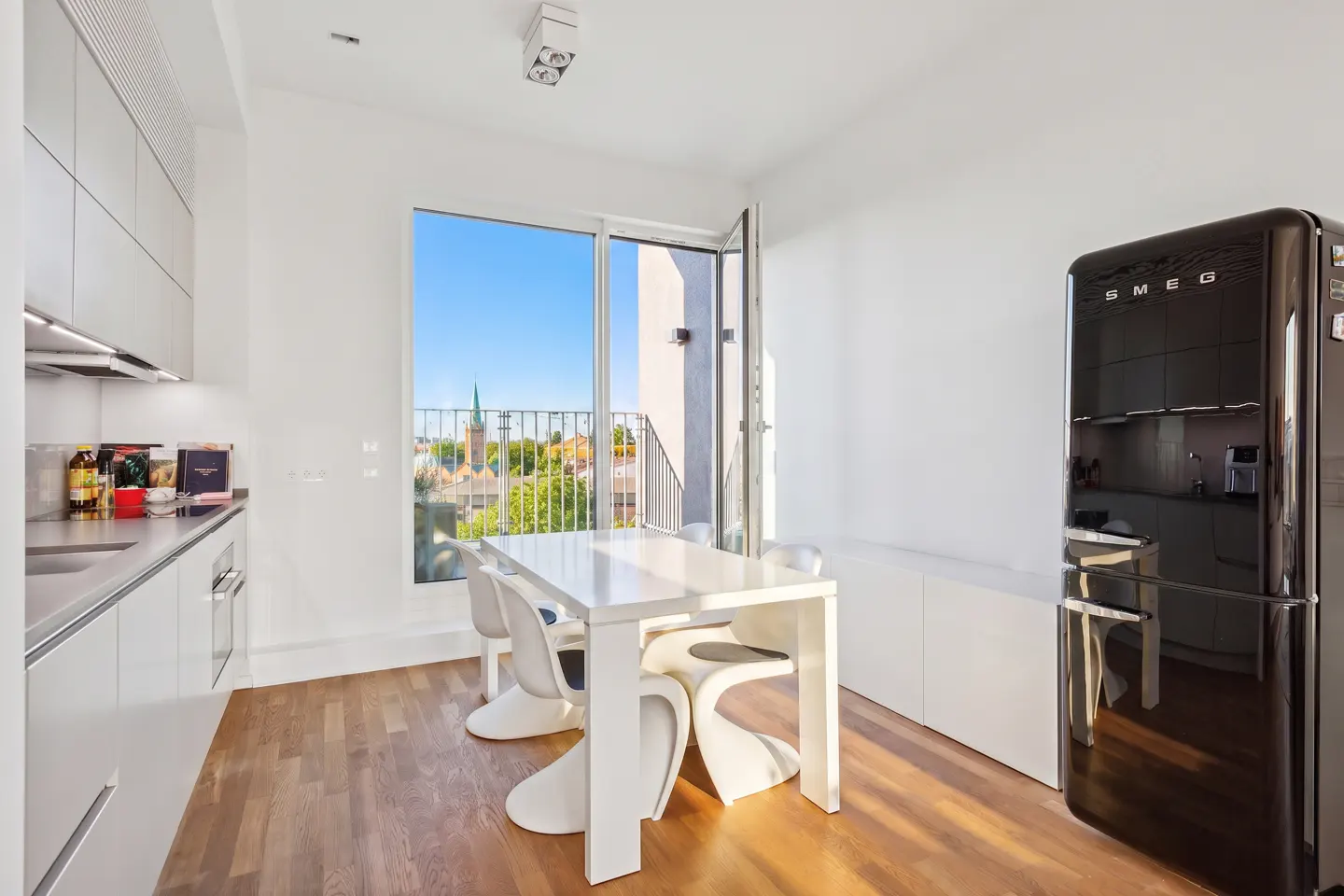 Bright kitchen with white cabinets, table, and chairs. A black SMEG refrigerator stands against the wall. Balcony doors open to a city view.
