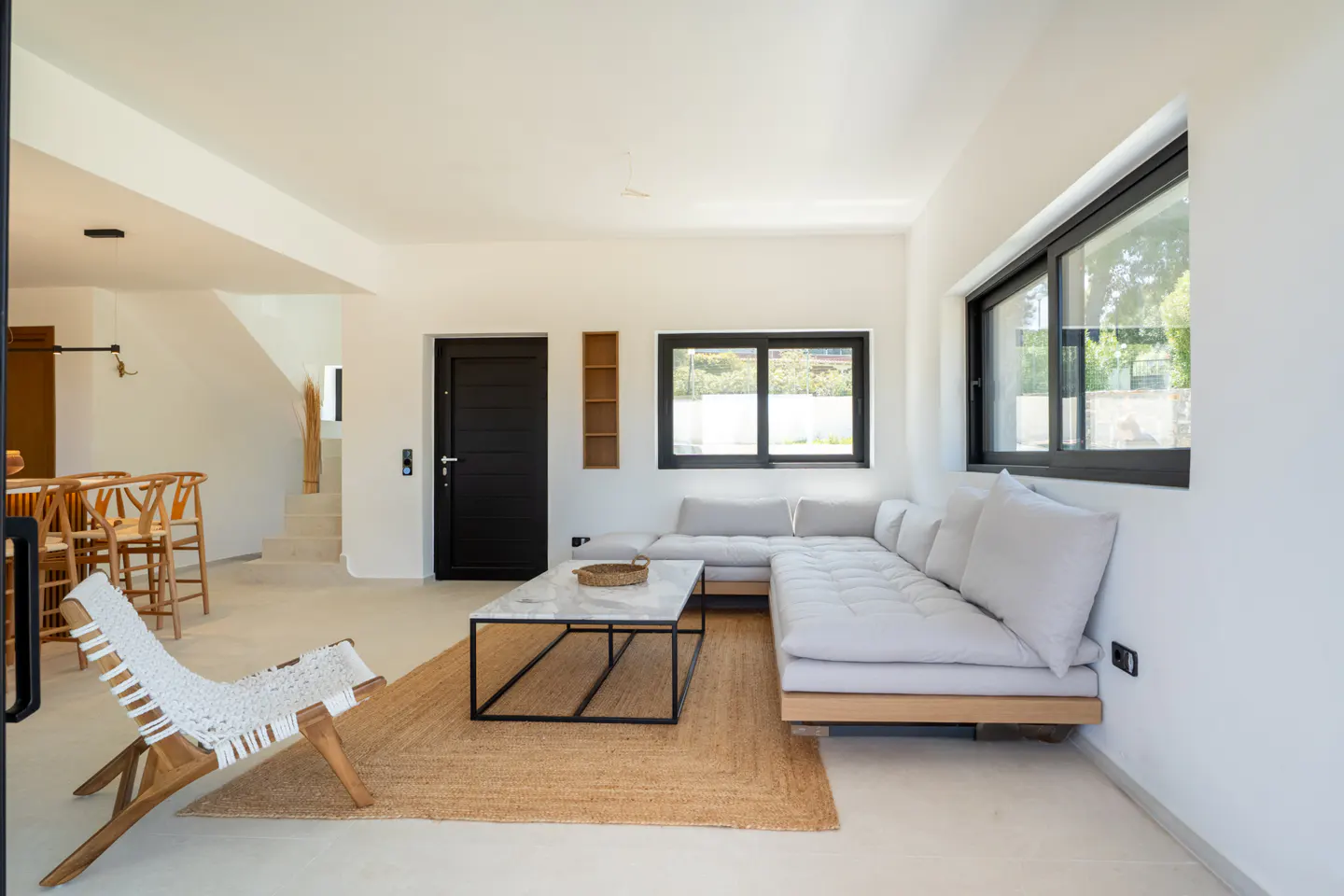 Bright living room with white walls, a gray sectional sofa, and a black-framed marble coffee table on a jute rug. A white woven chair sits near a black door.