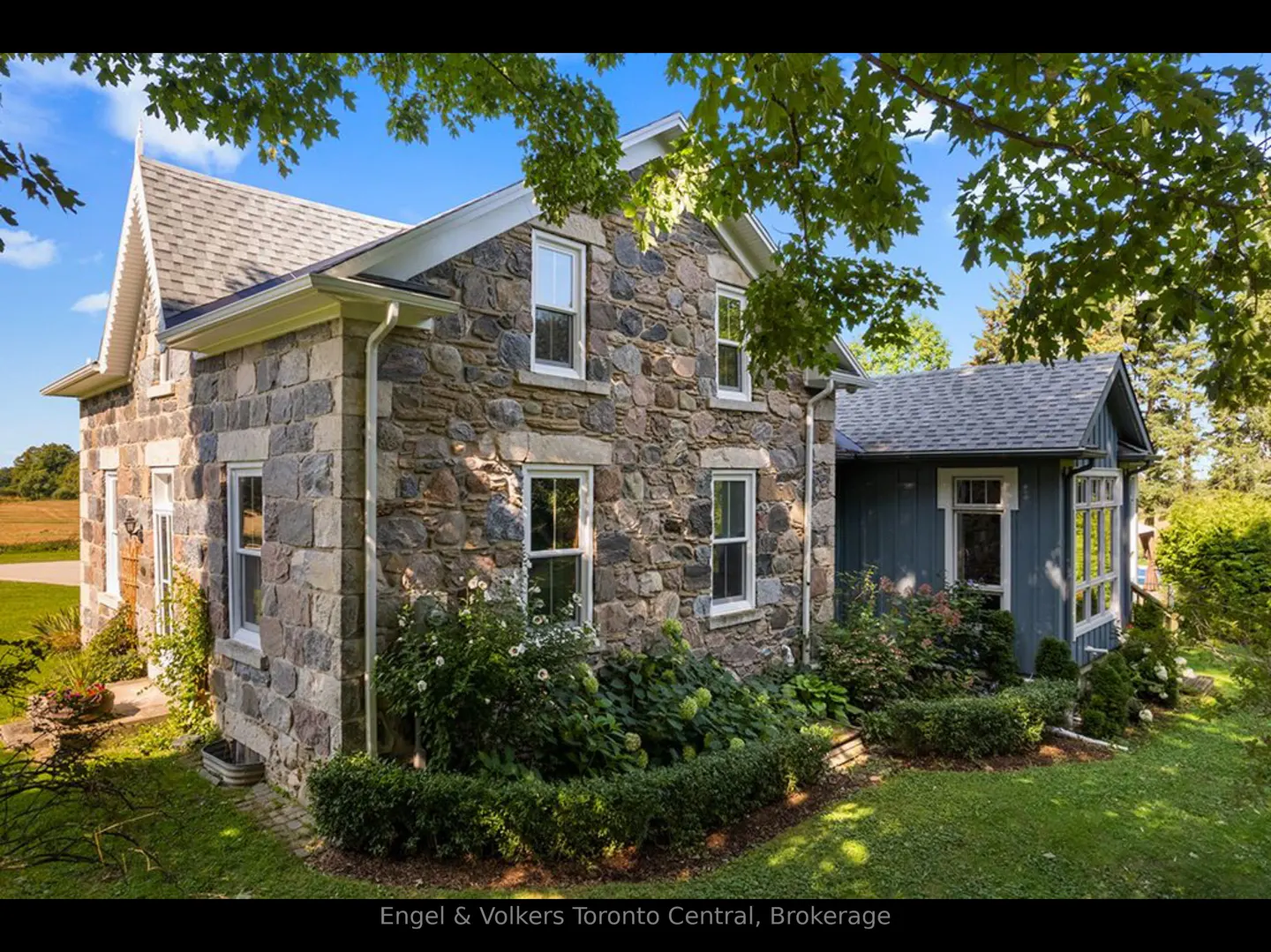 Exterior view of a two-story stone house with white-framed windows, a gray roof, and green landscaping. A blue addition is on the right.