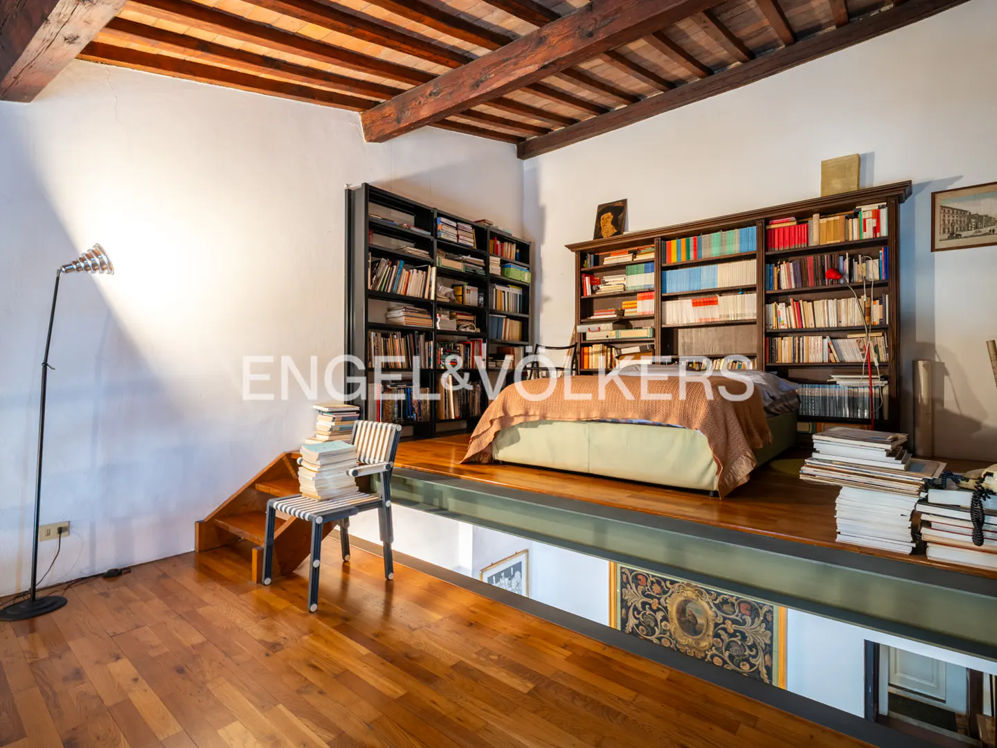 Interior view of a lofted bedroom with wood floors, exposed beams, and bookshelves. A chair with books sits near stairs leading to the platform bed.