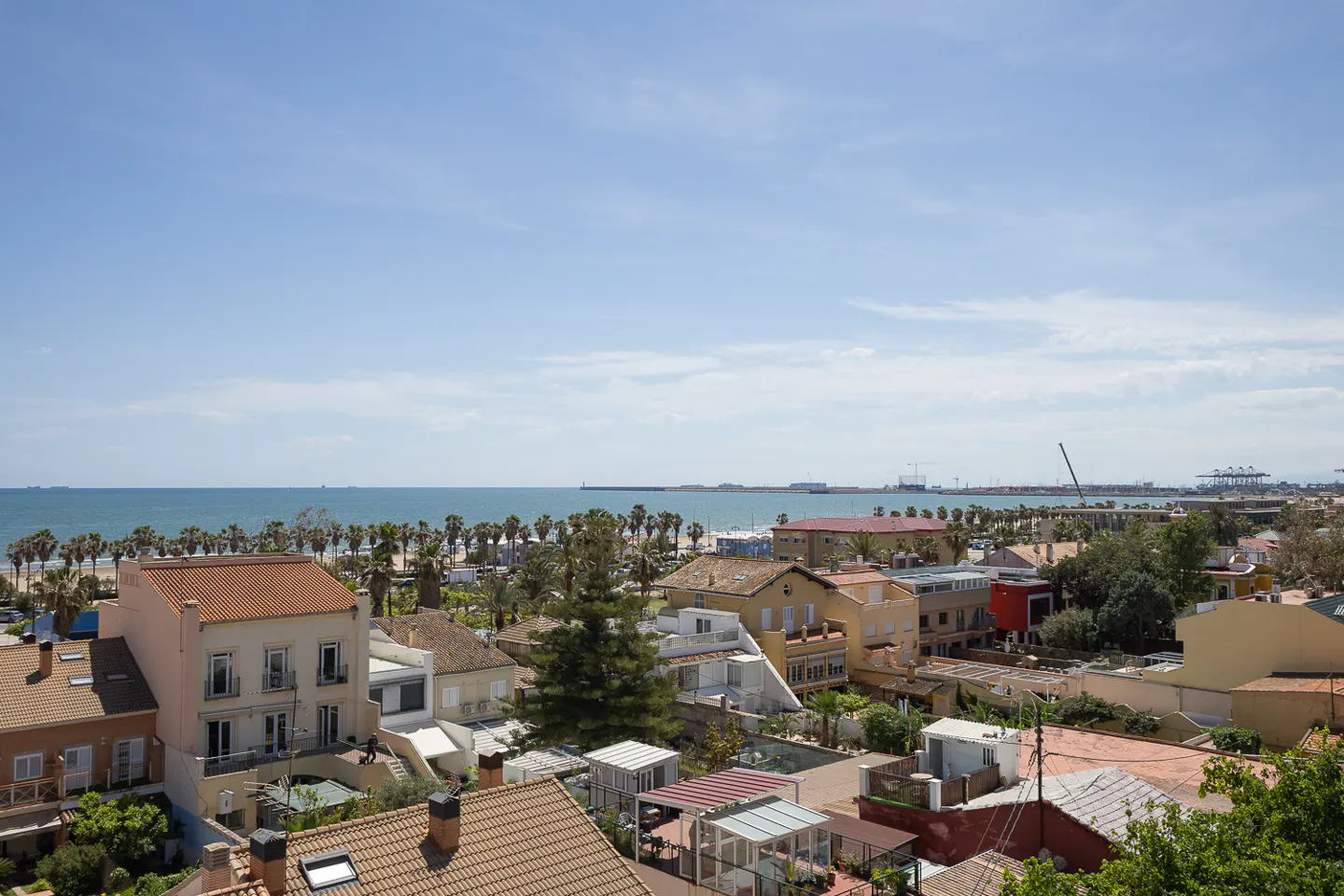 View of a coastal town with red-roofed buildings, palm trees, and the blue ocean under a bright sky.