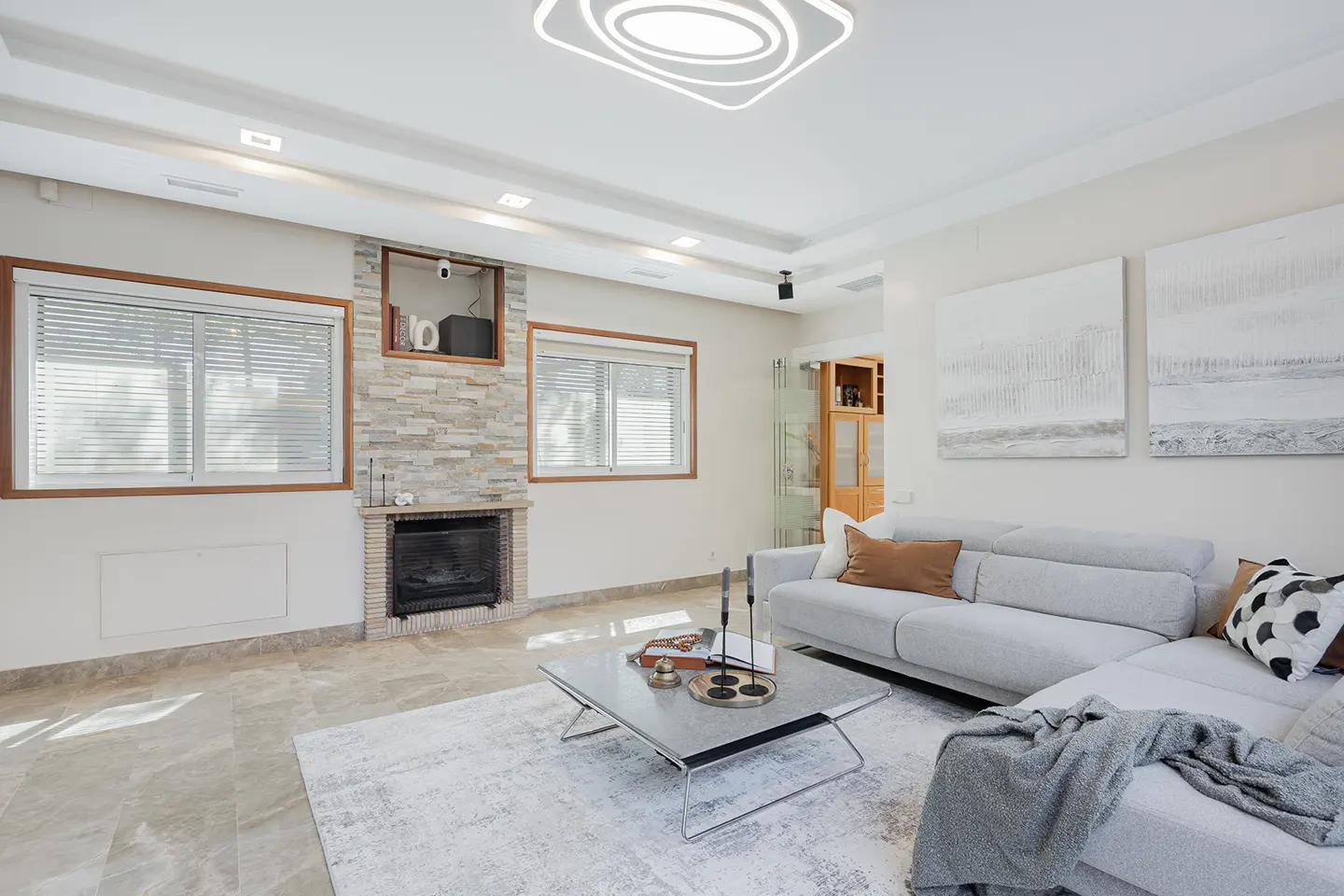 Bright living room with a gray sectional sofa, a stone fireplace, and a modern coffee table on a light rug. Two windows with blinds let in natural light.