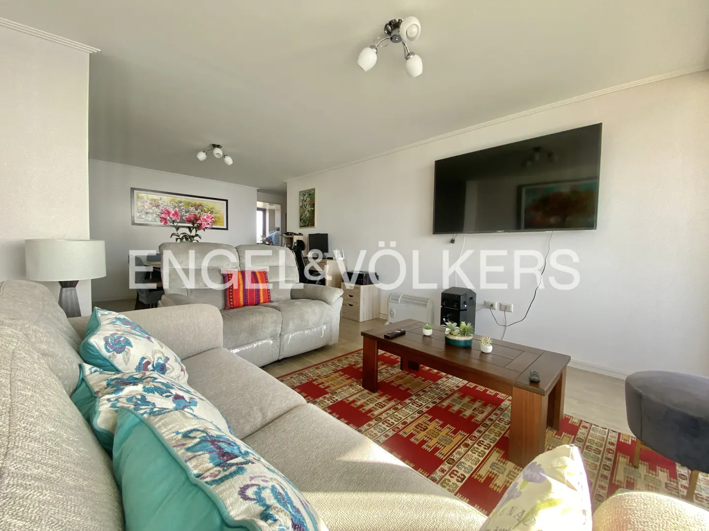 Living room with gray sofas, patterned pillows, a red rug, and a dark wood coffee table. A large TV hangs on the white wall.