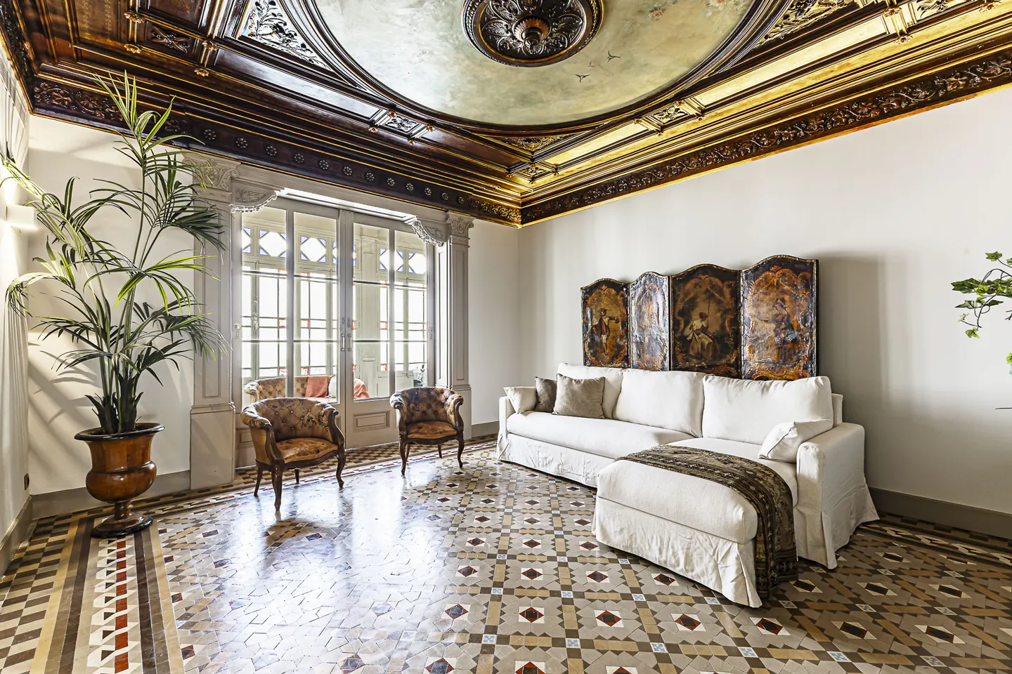 A bright living room with a white sofa, patterned chairs, and a decorative screen. The room features an ornate ceiling and tiled floor.