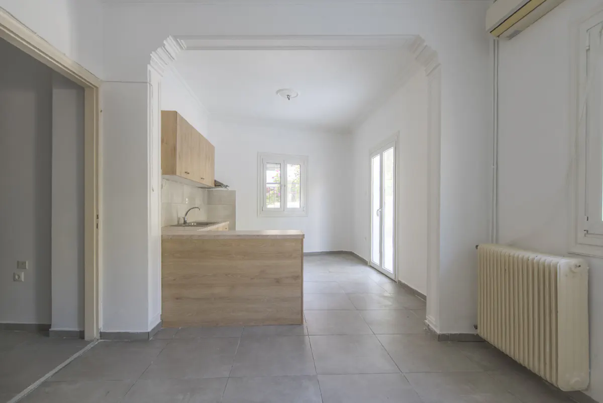 A bright, empty apartment with gray tile floors, white walls, and a light wood kitchen area. A doorway leads to another room.